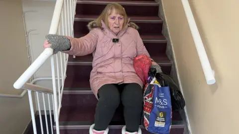 An elderly woman wearing a pink coat and pink boots is sat on a flight of stairs looking out of breath, carrying bags of shopping.