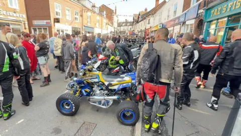 The picture shows a large crowd of about 50 people. They are all walking through a town centre, with shops either side of the photo. Down the middle of the crows are an array of motorbikes, which people are looking at. 