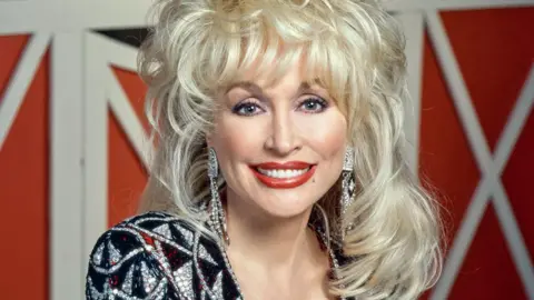 Getty Images/CBS photo Archive A head and shoulders shot. Dolly's blonde hair stands up in big waves and falls onto her shoulders. She wears a sliver and black diamante V neck dress with red accent colours. Long diamante earrings hang down.