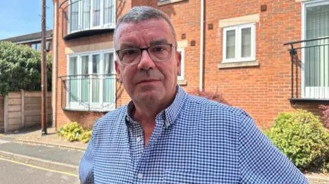 BBC/Charlotte Benton A man standing on a residential street with a red brick building behind him. He is wearing a blue and white check shirt and has short grey hair. He is wearing glasses and looking into the camera with a slight smile. 
