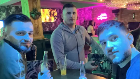 Greater Manchester Police Three men in a neon-lit bar smiling next to drinks.