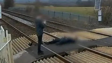 A blurred CCTV image shows a boy or man standing up taking a photo of another boy or man who is lying down on railway tracks at a level crossing. Their faces are blurred on purpose.