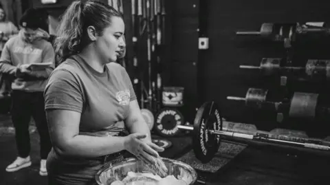 Gordon Siegal A black‑and‑white image shows Lauri standing beside a large bowl filled with chalk, rubbing hands in preparation for lifting. A loaded barbell rests on a platform nearby, with weight plates and racks visible against a dark wall in the background. Another individual stands behind holding a notepad, while various strength‑training equipment surrounds the area.