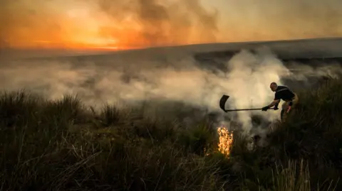 PA Media A firefighter is tackling a large amount of smoke in area which consists of long grass, the sunset and hills can be seen in the distance.