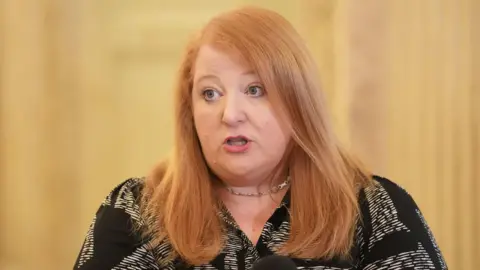 PA Media Naomi Long speaking. She has long ginger hair and is wearing a black and white blouse and a silver necklace.