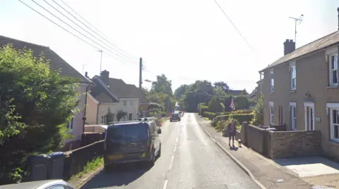 Google A general view of the road through Dennington. Houses line either side of the road as vehicles can be seen travelling down. On the right is a woman walking her dog along a pavement. 