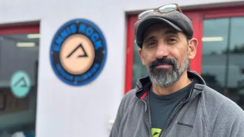 A man with a short black and grey beard wears a grey flat cap with glasses rested upon it. Behind him is a white building with a glass door with a red frame. There is also a window with a red frame. A blue sign sits between the two glass panels. 