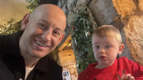 A family photo of Grant and Callum. Grant has a bald head and black jacket. Callum has short blond hair and wears a red sweater