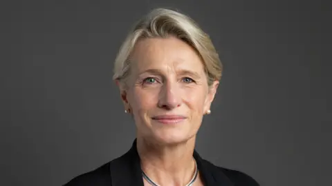 Alison Griffiths, a woman with with short and neat blonde hair, is pictured in an official Parliamentary portrait. She is smiling slightly and wearing a black jacket, necklace and pearl earrings. 