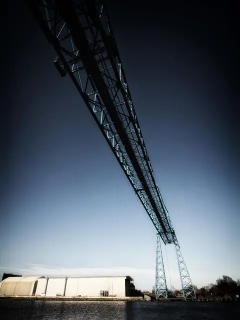 Geoff Ruffell A blue metal work bridge on tall legs straddles a river. A warehouse can be seen on the far bank and the photographer is looking up at the bridge.