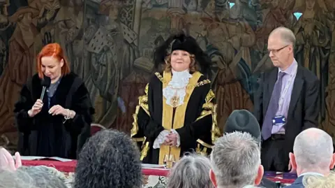 Rachel Lancaster stands between two people while dressed in the mayoral robes.