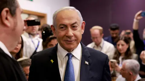 Netanyahu stands in a courtroom wearing a black suit jacket, a white shirt and a blue tie. Behind him the room is crowded, with several people holding their phones aloft to take photos of him. 