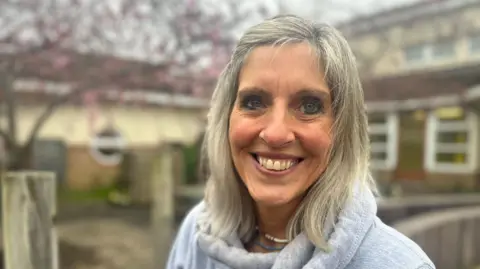 Aimee Dexter/BBC Sally Nye has short blonde hair. She is smiling and is wearing a grey jumper and a blue and white necklace. She is sitting in front of a building which is blurred in the background.