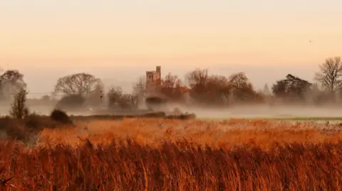 Amanda Norfolk A church sits on the horizon shrouded in mist. There are also trees either side of the church. In the foreground, there is a field of wheat almost orange in the early morning light.