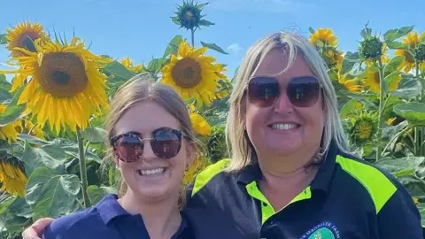 A head and shoulders image of two women wearing sunglasses while standing in front of a sunflower field. 