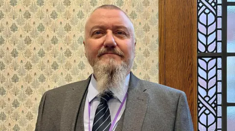 Councillor James Bean. He is looking at the camera. He has short hair, a long grey beard. He is wearing a light blue shirt, a dark blue tie and a grey jacket. 