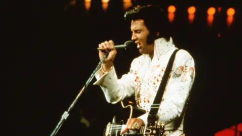 Getty Images Elvis Presley is pictured on stage in 1974 wearing his iconic white outfit and dark hair spruced up. He is singing into a mic, with his acoustic guitar hanging around him by a strap.