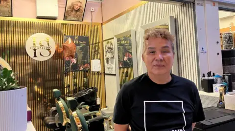 A far east asian man with short curly light brown hair is looking at the camera. He is wearing a black t-shirt with a white line in the shape of a cube. He is standing at the entrance to a salon area, behind him are two green barber chairs.