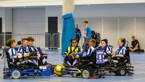 The Albion Foundation The West Bromwich Albion powerchair football team in a huddle. Six players are on their chairs and surrounding their coach. There is a ball in the middle of them.