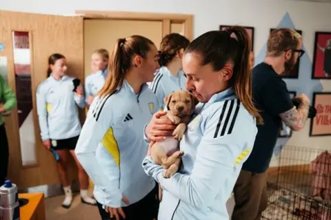 A woman in the foreground of the picture has her hair styled in a high ponytail and is dressed in light blue sports gear. She is holding a six-week old puppy and looking down at it. In the background there are four women who are also dressed in sporting clothes and a man in a black tshirt, glasses with tattoos on his arm appears to be holding a camera and looking to the right of the frame. 