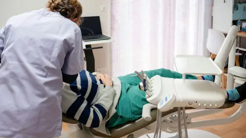Getty Images Shows a man in a dentist's chair, wearing a blue and grey striped jumper with green trousers. The female dentist has her back to the camera and is wearing a white dentist's gown. 