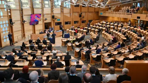 A wide shot of the Scottish Parliament chamber, with MSPs sitting at wooden desks in a wood-panelled room 