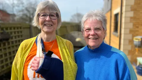 Two older women, perhaps in their 70s, smile as they stand in a back garden in a suburban home. The one on the left is a slightly taller lady with a grey bob, blue rimmed glasses, a mustard yellow cardigan, an orange and white T-shirt, and an arm in a navy sling. On the right is a lady with short grey hair, glasses with a light rim, and a bright blue turtleneck jumper. The sun is shining.