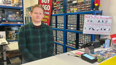 A man in a green checkered shirt is standing behind the counter of a shop. He has short ginger hair and a black t-shirt beneath his shirt. On the counter is a card machine, leaflets, till and receipt printer. Above the counter is a whiteboard with Lego figures taped to it and the handwritten title, "Hard To Find Minifigures." Behind the man are rows of shelves with small plastic transparent drawers, all labelled and filled with Lego bricks. Another shelving unit displays full Lego kits. There is also a red advertising flag behind him.