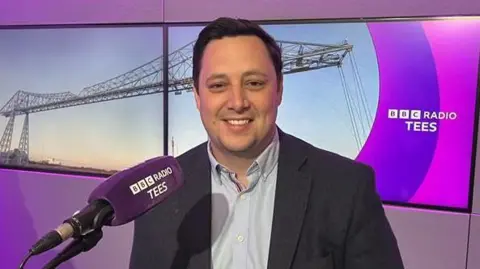 Ben Houchen sat in the BBC Radio Tees studio with a purple mic in front of him and two screens behind him with BBC Radio Tees written on them. He is smiling at the camera in a shirt and jacket.
