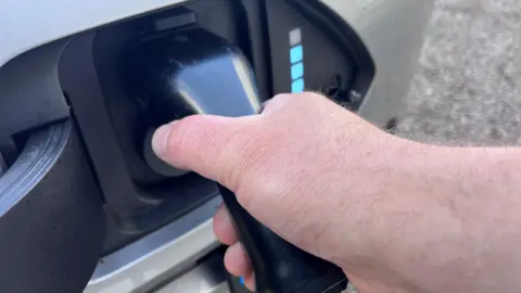 A right hand inserts a charger into the plug on an electric vehicle