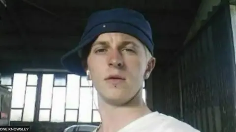 One Knowsley Michael Causer in a dark building with a panel of windows. He's wearing a white t-shirt and blue cap on sideways, with one headphone in