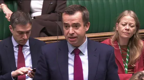 PPRIEN TV Calum Miller MP stands up to speak in Parliament. He had dark hair and was wearing a dark suit, a light and magenta shirt.