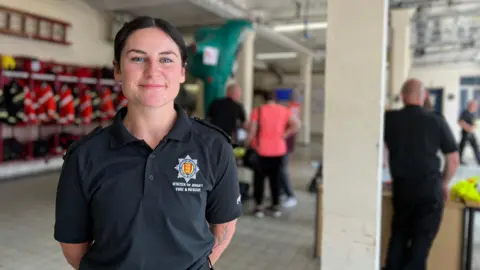 Beth smiles at the camera while she holds her hands behind her back. She is wearing a navy blue firefighter polo t-shire and has black hair tied into a ponytail. She is in the fire station and behind her firefighters are showing people equipment and their firefighting cloths are hung up on a rail.