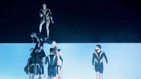 Circa tour video A tower of acrobats, supported by six performers on the left side of the frame. Men and women hold two female acrobats, one on her back is supported by her arms and legs, while the other stands on her torso looking out to the crowd. To the right of frame, a man looks at them across the stage.