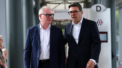 Two men in dark suits, open-necked white shirts and glasses walk through a modern bus station while talking to each other. On the left, Richard Parker has greying fair hair. On the right, Andy Burnham has dark hair with his arm at Parker's back. In the background, a bus stop branded with a West Midlands Travel logo is visible. 