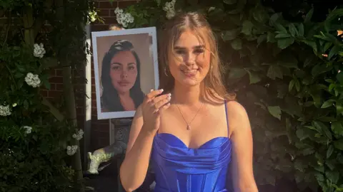 Family handout Delsie is wearing a cobalt blue evening gown and has her light brown hair down but off her face. She is smiling and holding a picture of her mum.