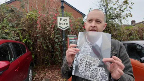 Richard Horton holding a see through A4 sleeve full of QR code stickers in a car park. Behind him are two cars and a ticket machine with a 'Pay here' sign above it. 