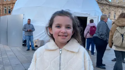Chloe has long brown hair and is wearing a cream-coloured, fuzzy zip‑up jacket. She stands smiling outdoors in front of a large white dome structure. Several adults are walking or standing in the background on a paved square, with historic buildings visible behind the dome.