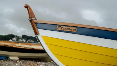 Kirstin McEwan A close up of the wooden hull of a rowing boat. It is painted blue, white and yellow. It has the name "Stranraer". Another boat is on the shore in the background.