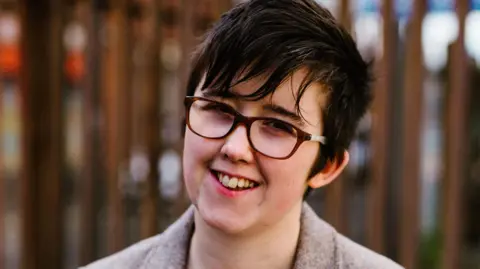Lyra McKee, a woman with short dark hair, smiling at the camera with her head slightly tilted to one side. She is wearing brown-rimmed glasses and a beige tweed blazer. She is standing in front of a tall steel security fence. 