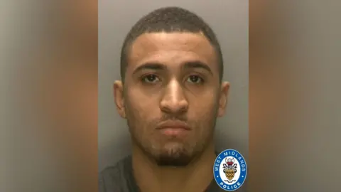 West Midlands Police Police mugshot of a man with closed-cropped dark hair, trimmed beard and stubble. He is wearing a grey T-shirt and standing against a grey background.