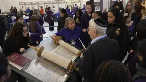 A group of school pupils stand around a table with large open scrolls, listening to an older man speaking.
