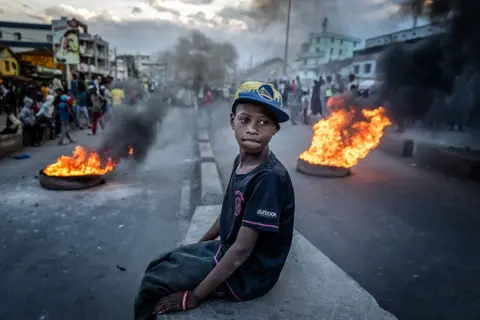 LUIS TATO / AFP / GETTY IMAGES A boy sits on a traffic wall as tyres burn either side in the background. His expression is inquisitive.