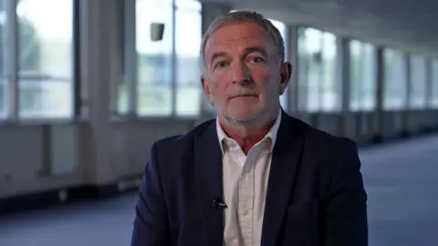 Andrew wearing a dark blazer and light shirt, seated indoors in a modern office corridor with large windows and soft natural light in the background.