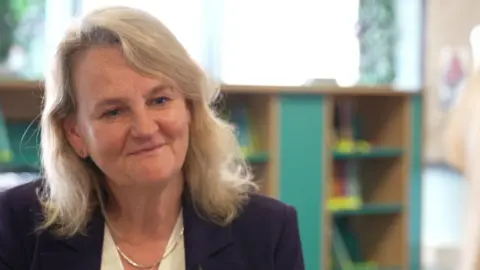 Lib Peck has blonde, shoulder length hair and is wearing a purple suit jacket. She is sitting in a classroom in front of a bookshelf
