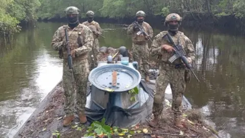 Four armed and uniformed members of the Ecuadoran military stand on top of a semi-submersible vessel in a mangrove-lined swamp.
