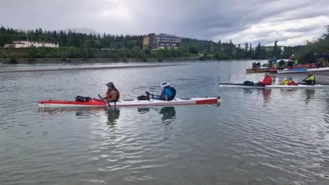 Jim Nye Ian Tyers and Jim Nye are in a canoe on the River Yukon. They are wearing life jackets and hats and there are several other canoes on the water behind them. There are several buildings in the distance and fir trees are growing near the far river bank.