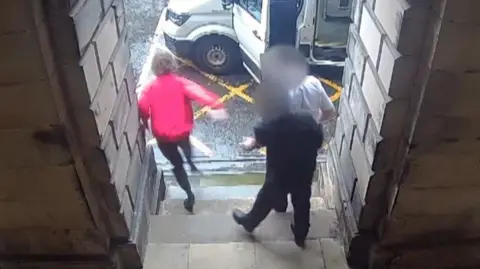 A person wearing a red jacket runs away from two men in uniforms outside a court building with a nearby van parked outside with its door open