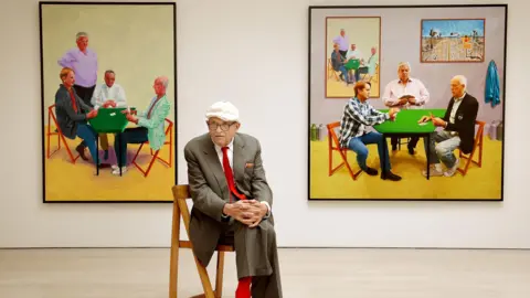 PA Media David Hockney sits in a brown suit with a red tie. He's in front of his work entitled Card Players (left) and A Bigger Card Players at the launch of an Exhibition of his work called David Hockney: Painting And Photography at Annely Juda Fine Art in central London.