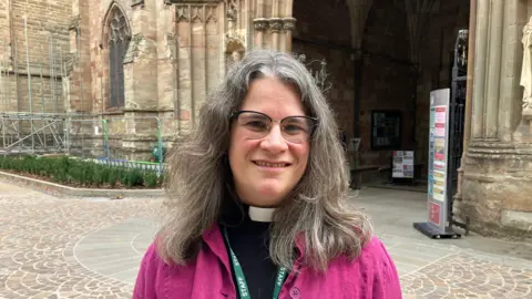 Canon Kimberly Bohan pictured outside the cathedral. She is wearing a pink overshirt over her traditional liturgical vestments. She has long grey hair and is wearing a pair of thin black glasses. 